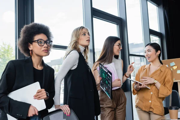 Young businesswoman pointing with finger while talking to asian colleague near multiethnic managers — Stock Photo