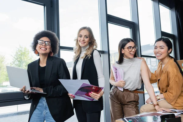 Happy multiethnic businesswomen with magazines and gadgets smiling at camera in office — Stock Photo