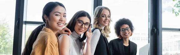 Giovane donna d'affari che regola gli occhiali mentre guarda la fotocamera con felici colleghi multietnici, banner — Foto stock