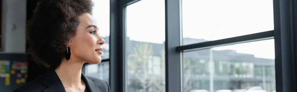 Vue de côté de femme d'affaires afro-américaine positive regardant par la fenêtre dans le bureau, bannière — Photo de stock