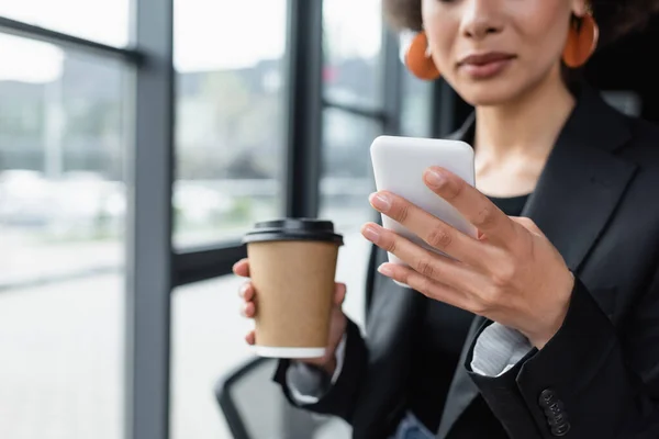 Обрезанный вид размытой африканской бизнесвумен с бумажной чашкой чата на сотовый телефон в офисе — стоковое фото