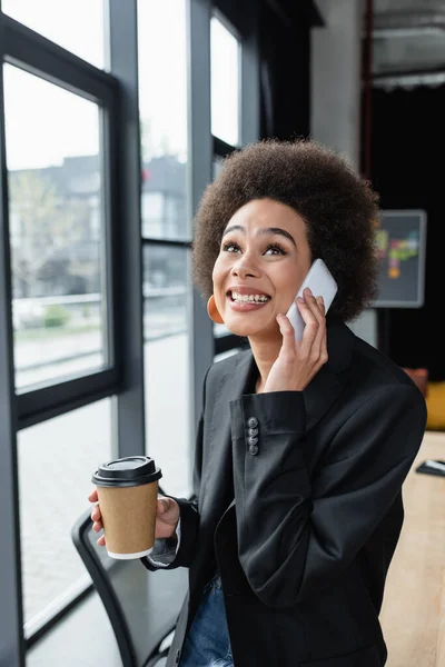 Emocionada mujer de negocios afroamericana con bebida para llevar hablando por teléfono móvil en la oficina - foto de stock