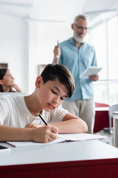 Asian schoolboy writing on notebook near blurred teacher — Stock Photo