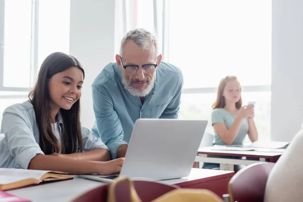 Teacher looking at laptop near smiling schoolgirl in classroom — Stock Photo