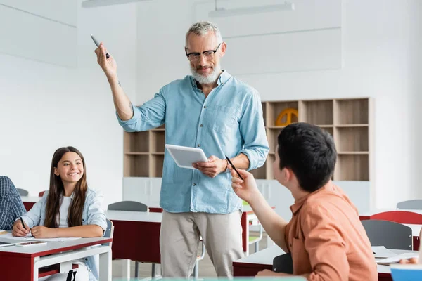 Profesor con marcador y tableta digital de pie cerca de los alumnos - foto de stock