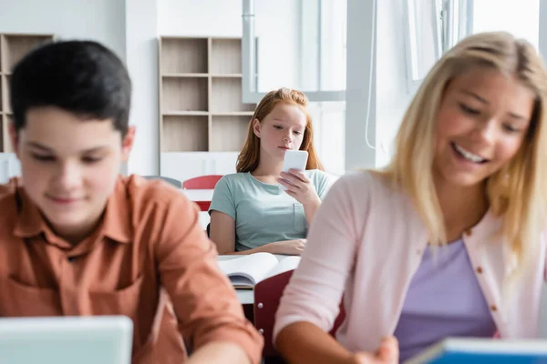 Colegiala usando teléfono celular cerca de compañeros de clase borrosos en el aula - foto de stock