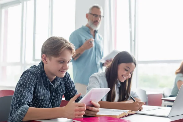 Écolier souriant utilisant une tablette numérique près d'une fille écrivant sur un cahier et enseignant flou — Photo de stock