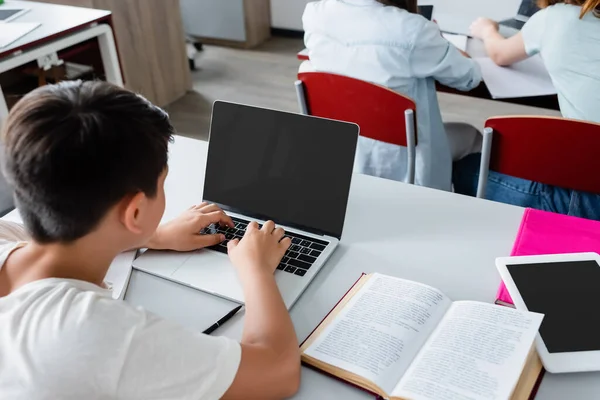 Vista aérea del colegial usando el ordenador portátil con la pantalla en blanco cerca del libro - foto de stock