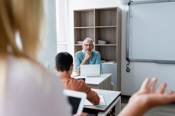 Smiling teacher looking at blurred pupil near laptop in classroom — Stock Photo