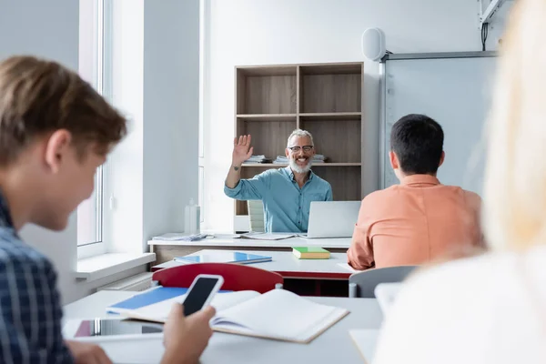 Professeur joyeux agitant la main aux élèves sur le premier plan flou en classe — Photo de stock