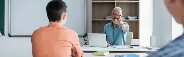 Cheerful teacher pointing with finger near blurred schoolkids in classroom, banner — Stock Photo