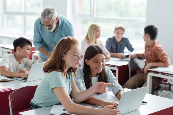 Colegial apuntando al portátil cerca del maestro y compañeros de clase multiétnicos - foto de stock