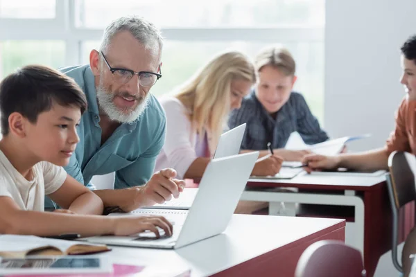 Mature teacher pointing at laptop near asian pupil in classroom — Stock Photo