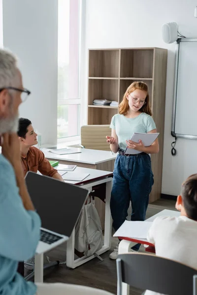 Schülerin schaut auf Notizbuch, während sie in der Nähe von Klassenkameraden und Lehrer spricht — Stockfoto