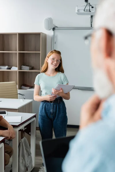 Une écolière souriante tenant un cahier près d'un professeur flou en classe — Photo de stock