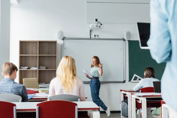 Smiling pupil with notebook talking near erase board and classmates — Stock Photo