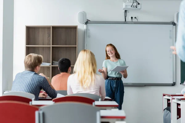 Escolar sorrindo com notebook falando perto apagar placa e colegas de classe — Fotografia de Stock