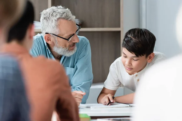 Mature teacher looking at asian schoolboy with notebook — Stock Photo