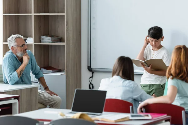 Asian schoolboy looking at notebook near erase board and teacher — Stock Photo