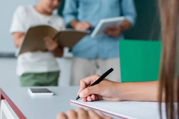 Vista recortada de los escolares escribiendo en un cuaderno cerca del teléfono inteligente en el aula - foto de stock