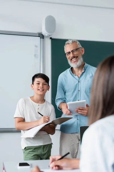 Sonriente profesor con digital tablet mirando a cámara cerca asiático alumno con notebook - foto de stock