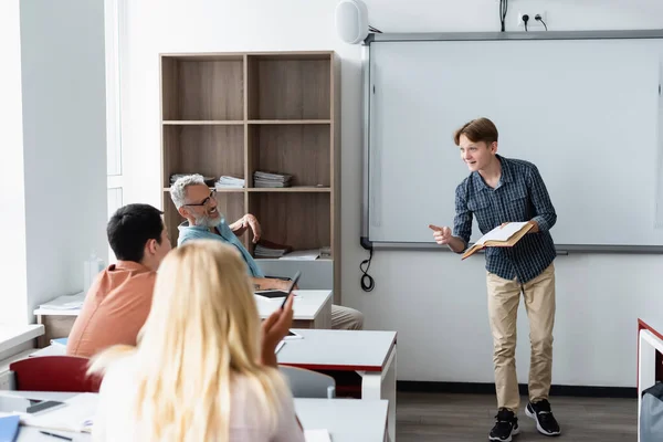 Pupilla sorridente con libro che punta con dito vicino a insegnante e compagni di classe — Foto stock