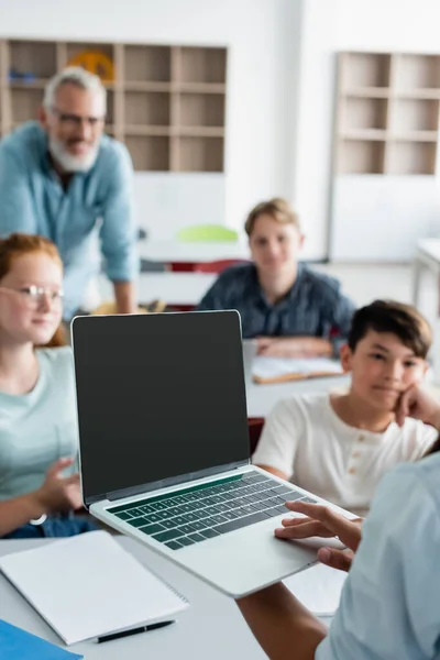 Colegial sosteniendo portátil cerca borrosa compañeros de clase multiétnicos y maestro - foto de stock