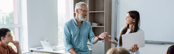 Écolière souriante avec ordinateur portable regardant un professeur mature en classe, bannière — Photo de stock