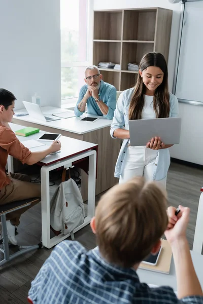 Lächelndes Schulmädchen mit Laptop in der Nähe von Lehrern und Schülern — Stockfoto