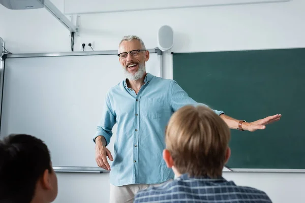 Lächelnder Lehrer spricht neben verschwommenen Schülern und Kreidetafel — Stockfoto