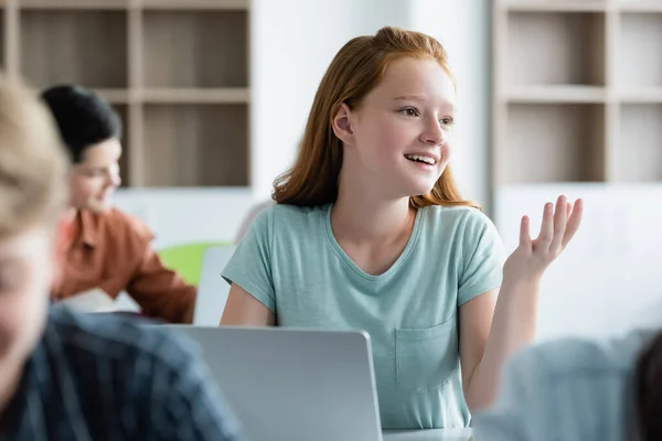 Escolar sonriente mirando hacia otro lado cerca de la computadora portátil y compañeros de clase borrosos - foto de stock