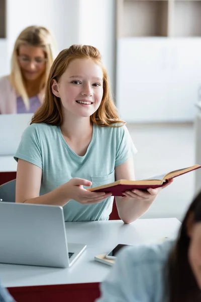 Smiling schoolgirl holding book near devices in classroom — Stock Photo