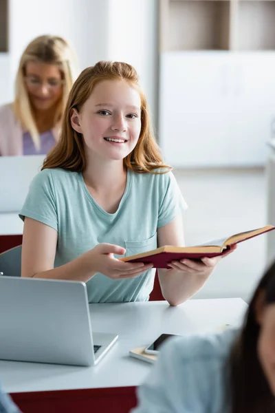 Colegiala sonriente sosteniendo libro cerca de dispositivos en el aula - foto de stock