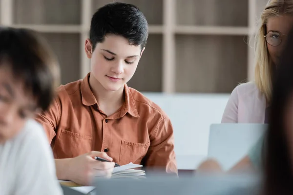 Schoolboy holding pen near notebook and blurred multiethnic pupils — Stock Photo