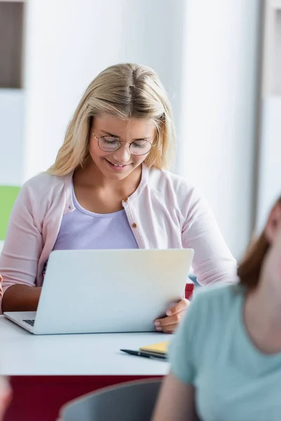 Smiling schoolgirl in eyeglasses using laptop during lesson in classroom — Stock Photo