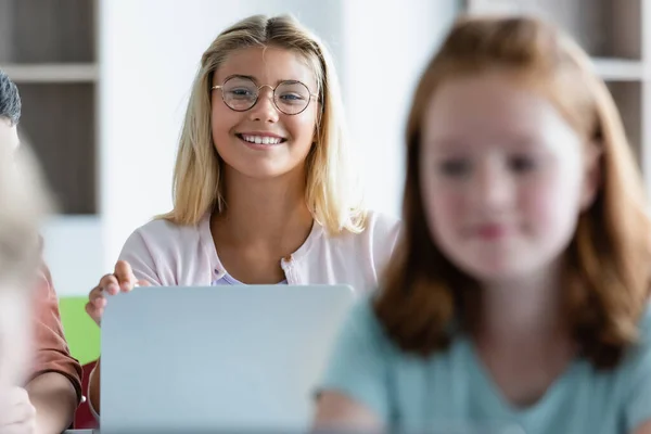 Chica alegre mirando a la cámara cerca de la computadora portátil y compañeros de clase - foto de stock