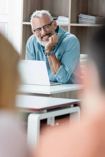 Lächelnd reifer Lehrer sitzt neben Laptop in der Schule — Stockfoto