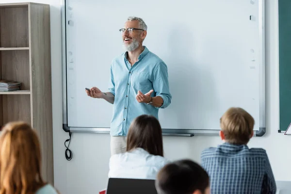 Profesora positiva con marcador hablando cerca del tablero de borrado y escolares en el aula - foto de stock