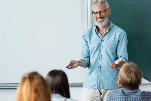 Cheerful mature teacher with marker pointing at blurred schoolkids near erase board — Stock Photo