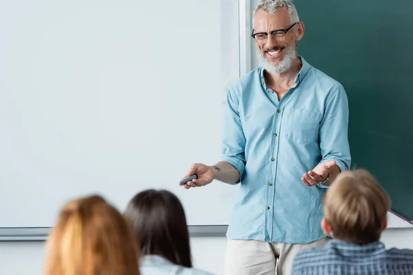 Fröhlich reifer Lehrer mit Marker, der auf verschwommene Schüler in der Nähe der Löschtafel zeigt — Stockfoto