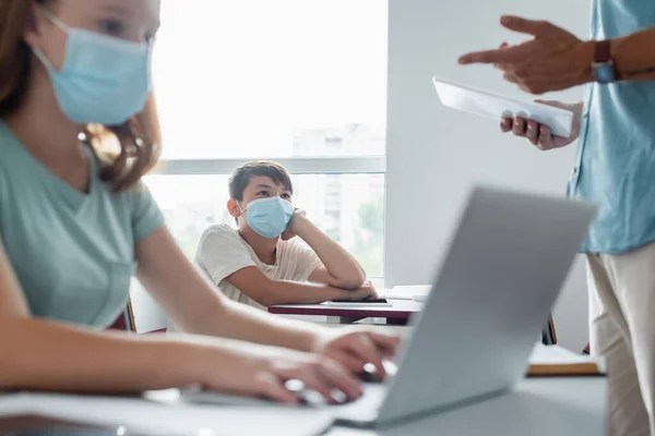 Asiáticos colegiales en máscara médica mirando borrosa profesor con tableta digital - foto de stock