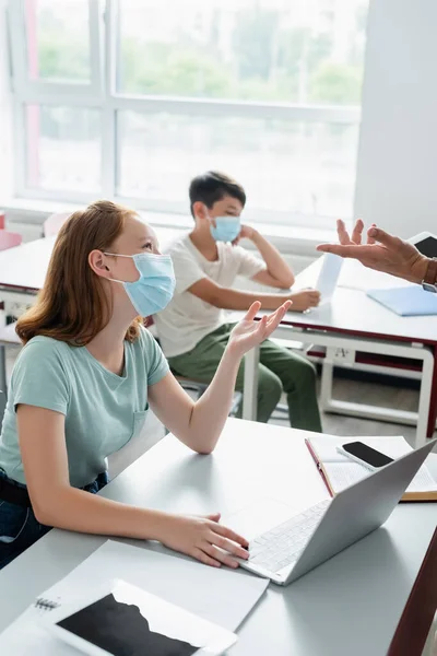Écolière en masque médical parlant à l'enseignant avec tablette numérique pendant la leçon — Photo de stock