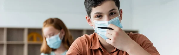 Школярка в медичній масці тримає ручку в класі, банер — стокове фото