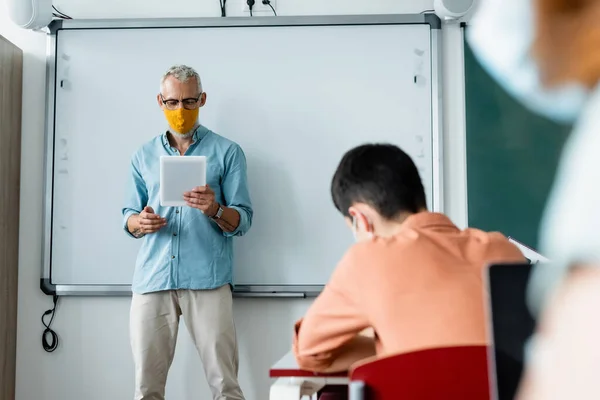 Учитель в защитной маске держит цифровой планшет рядом с размытыми зрачками во время урока — стоковое фото