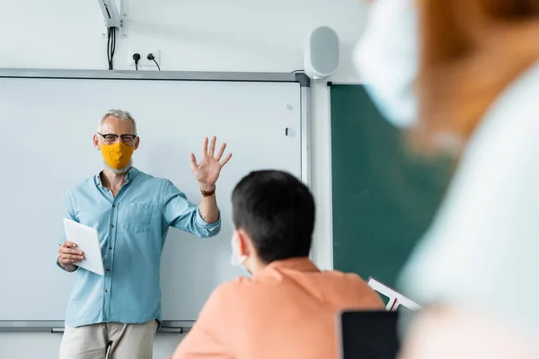 Учитель в защитной маске держит цифровой планшет и показывает ладонь рядом с учениками — стоковое фото