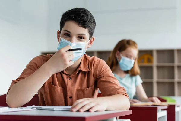 Écolier en masque médical tenant un stylo près d'un ordinateur portable et un ami flou — Photo de stock