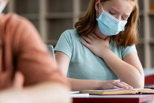 Malade écolière en masque médical assis près du livre — Photo de stock