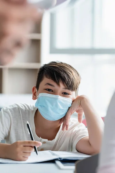 Азиатский школьник в медицинской маске держит ручку и смотрит в камеру — стоковое фото