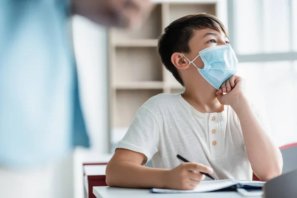 Азиатский школьник в медицинской маске держит ручку рядом с размытым учителем — стоковое фото