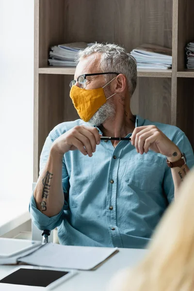 Teacher in protective mask holding pen near notebooks and digital tablets on table — Stock Photo